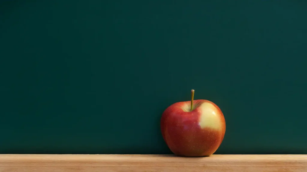 An apple sits on a wooden table in front of a green chalkboard.